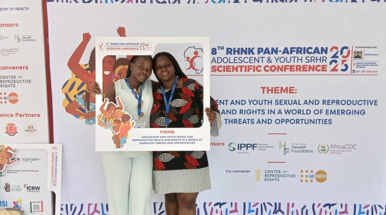 Make Way youth representatives Vilda Atieno and Elizabeth Warindi are standing with a Polaroid frame prop in front of a conference board depicting logos and the theme of the 8th Reproductive Health Network Kenya (RHNK) Adolescents and Youth Conference in Mombasa, Kenya.