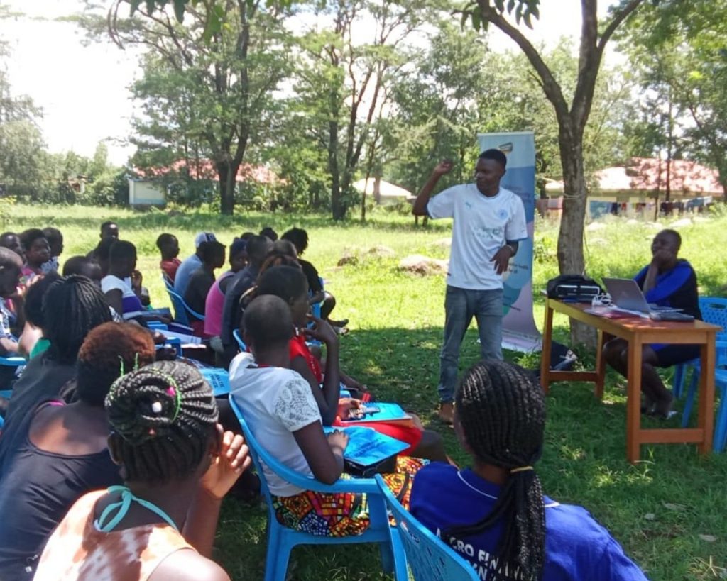 Sylvester Adhaya is standing and presenting in front of a group of youth in a green park area.