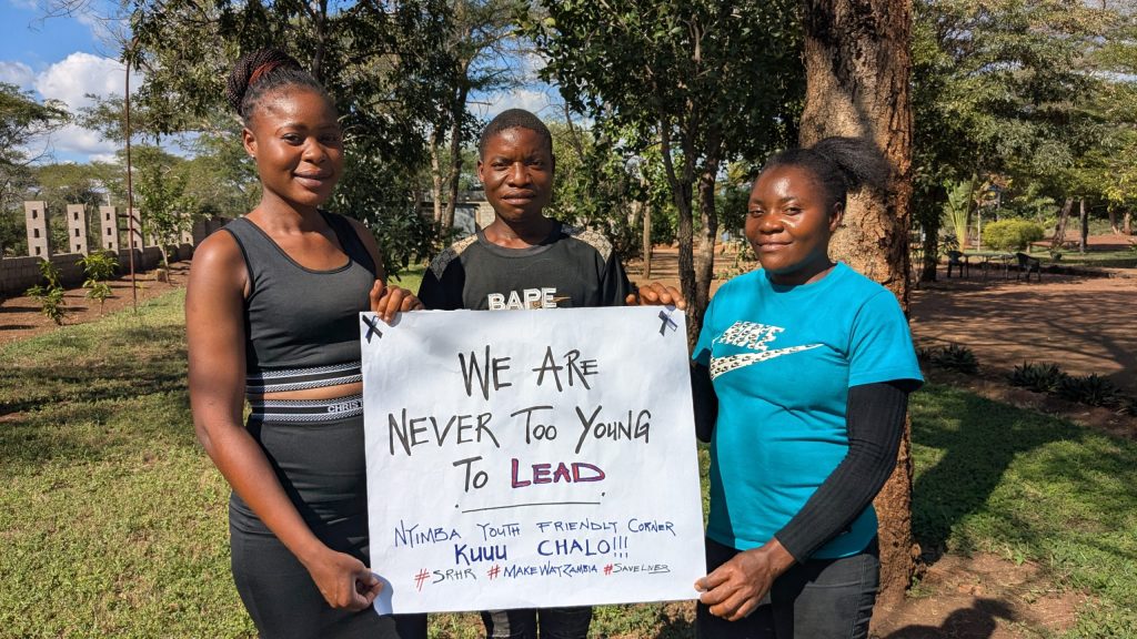 Three young people standing in a park with trees behind them, holding a sign that says: "We are never too young to lead. Nyimba Youth Friendly Corner. Kuuu Chalo!!! #SRHR #MakeWayZambia #SaveLives"