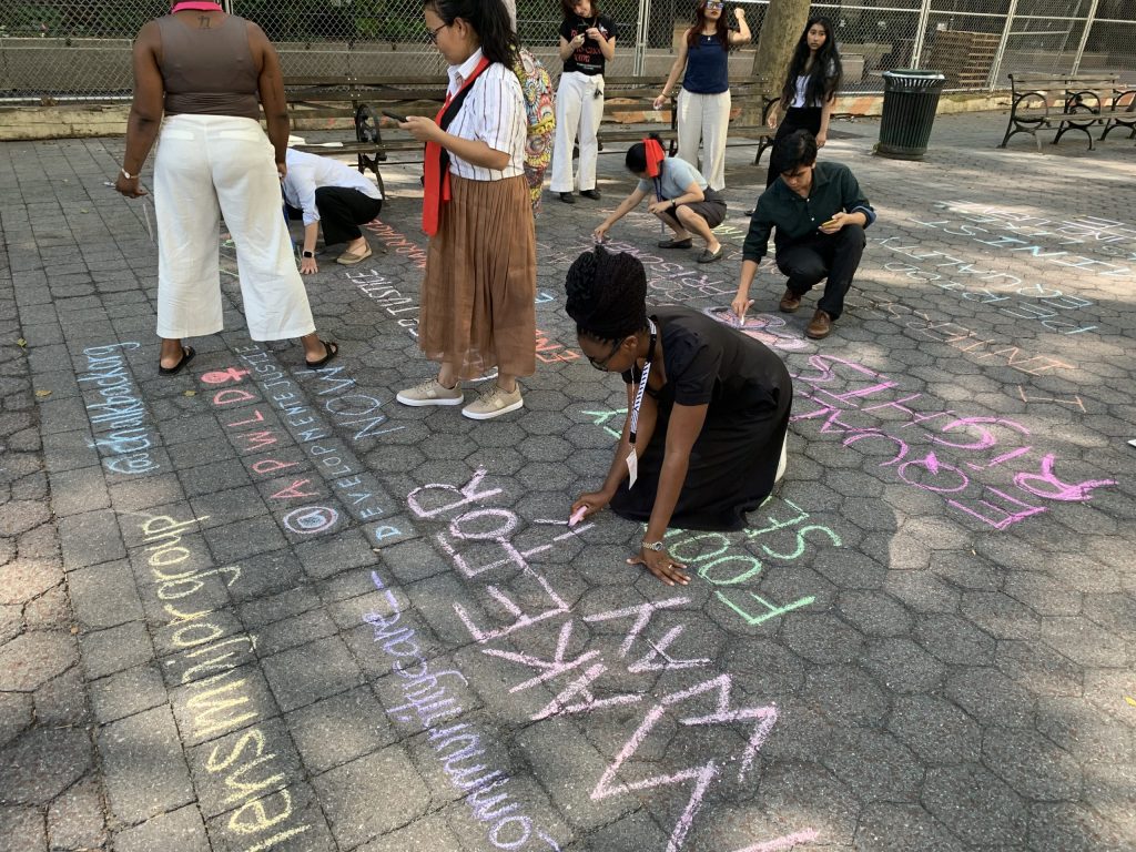 Bertha writing 'Make Way for youth' with chalk on the pavement with several other people around her.